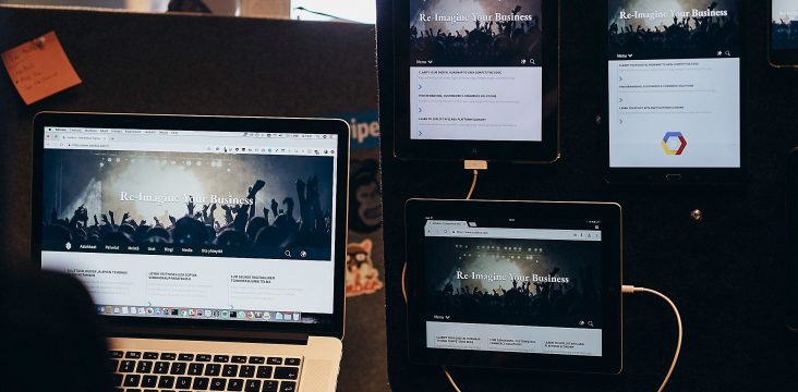 Tumma seinä, jolla tabletteja ja puhelimia latausjohdoissa, edessä läppäri – Miksi yritys tarvitsee verkkosivuston? Aloita tavoitteesta