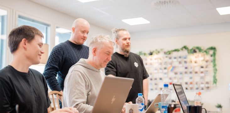 Neljä ihmistä läppärin ääressä, taustalla vihreällä köynnöksellä koristeltu valokuvaseinä – Nokia myytiin Microsoftille, mitä nyt Suomi?