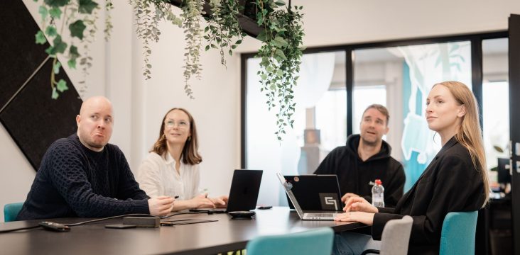 Pitkähiuksinen henkilö mustassa takissa istuu pöydän päässä muihin päin – Digitaalisen liiketoiminnan trendit 2022