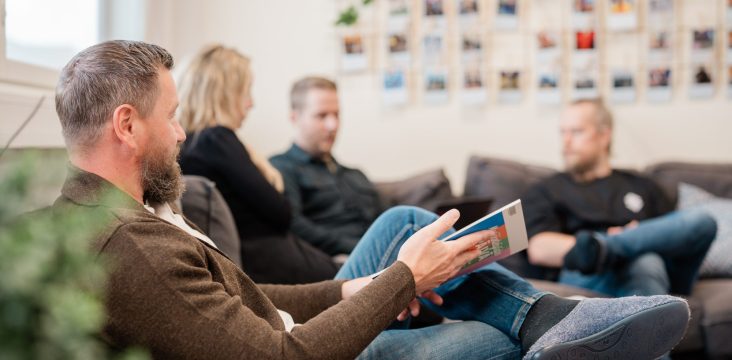 Parrakas henkilö ruskeassa takissa katsoo tablettia sohvalla, vieressä kollegoita – Miksi verkkokauppa pelottaa?