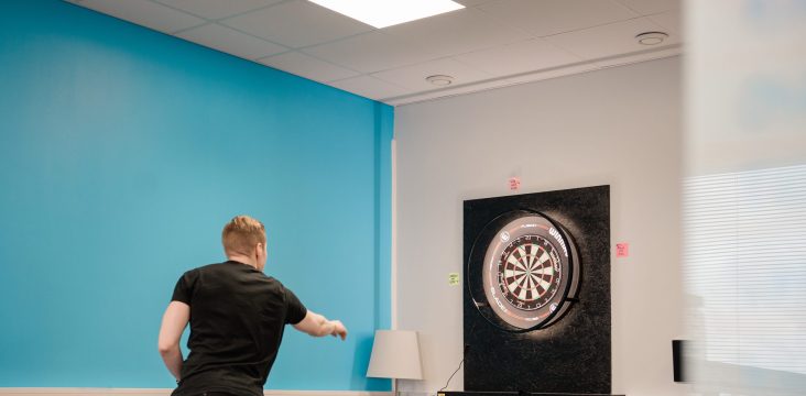 A dartboard and a floor lamp in the corner of a break room, next to a turquoise wall – Product-Led Vs Sales-Led: An Introduction To Product-Led Sales