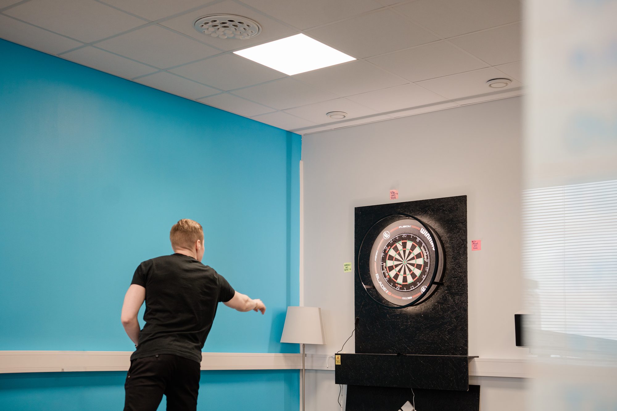 A dartboard and a floor lamp in the corner of a break room, next to a turquoise wall – Product-Led Vs Sales-Led: An Introduction To Product-Led Sales
