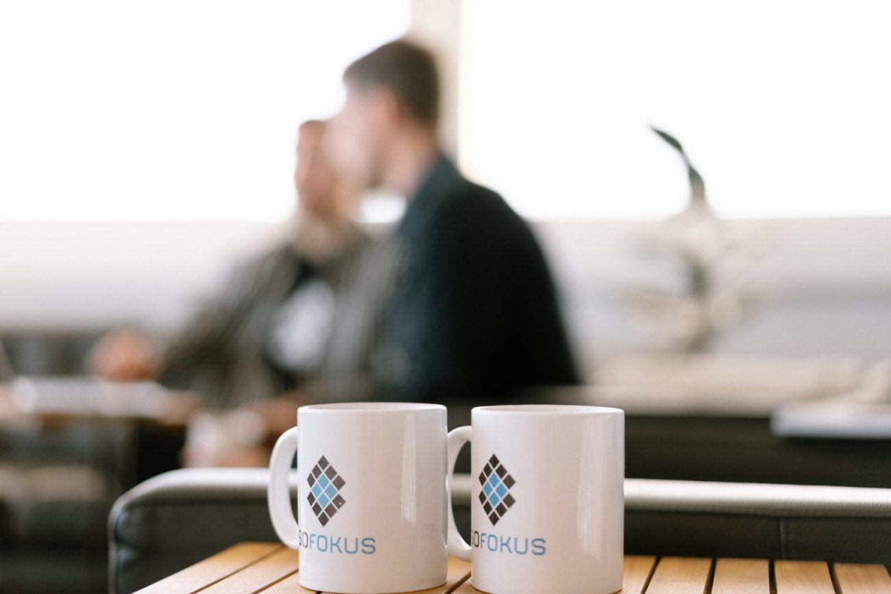 Two branded Sofokus coffee mugs on a table with colleagues meeting in the background