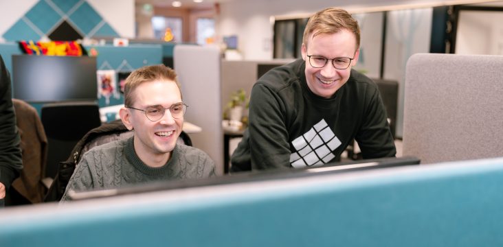 Two colleagues looking at a screen together and smiling, a teal diamond-patterned wall behind them – What does digital development take in the age of AI? Three capabilities that matter more than coding