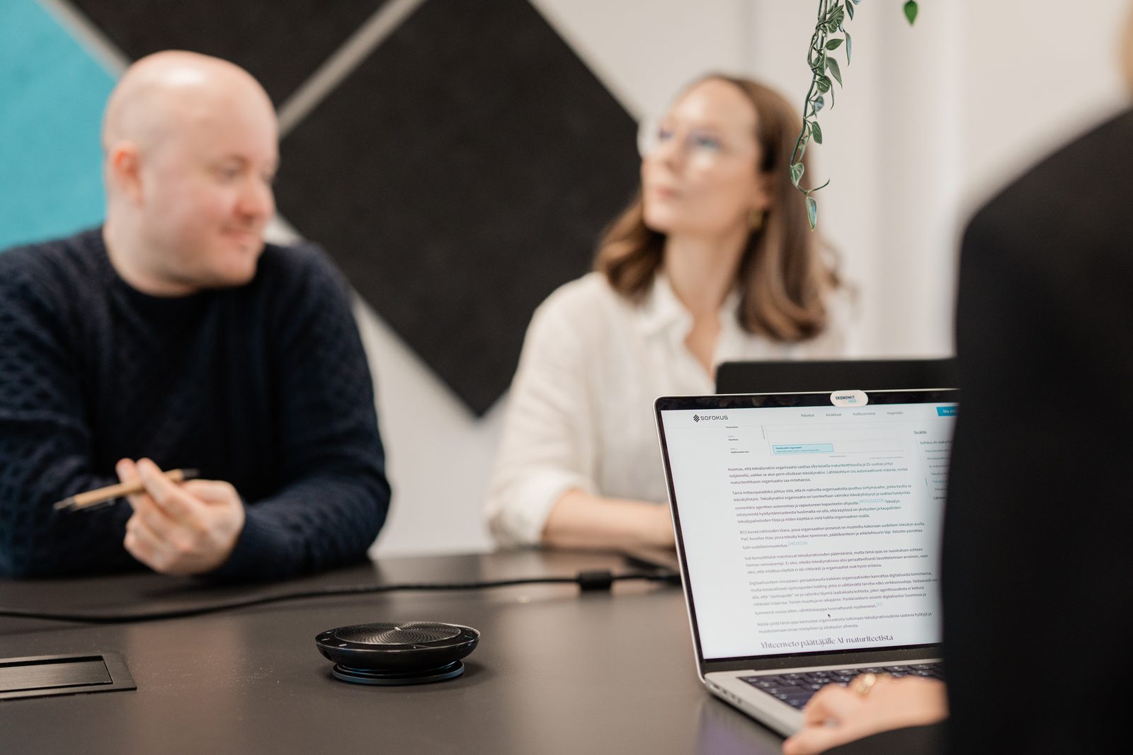 Kolme asiantuntijaa palaverissa, henkilö pitää kynää kädessään ja etualalla on kannettava, jonka näytöllä on dokumentti / Three experts in a meeting, a person holding a pen, with a laptop showing a document in the foreground