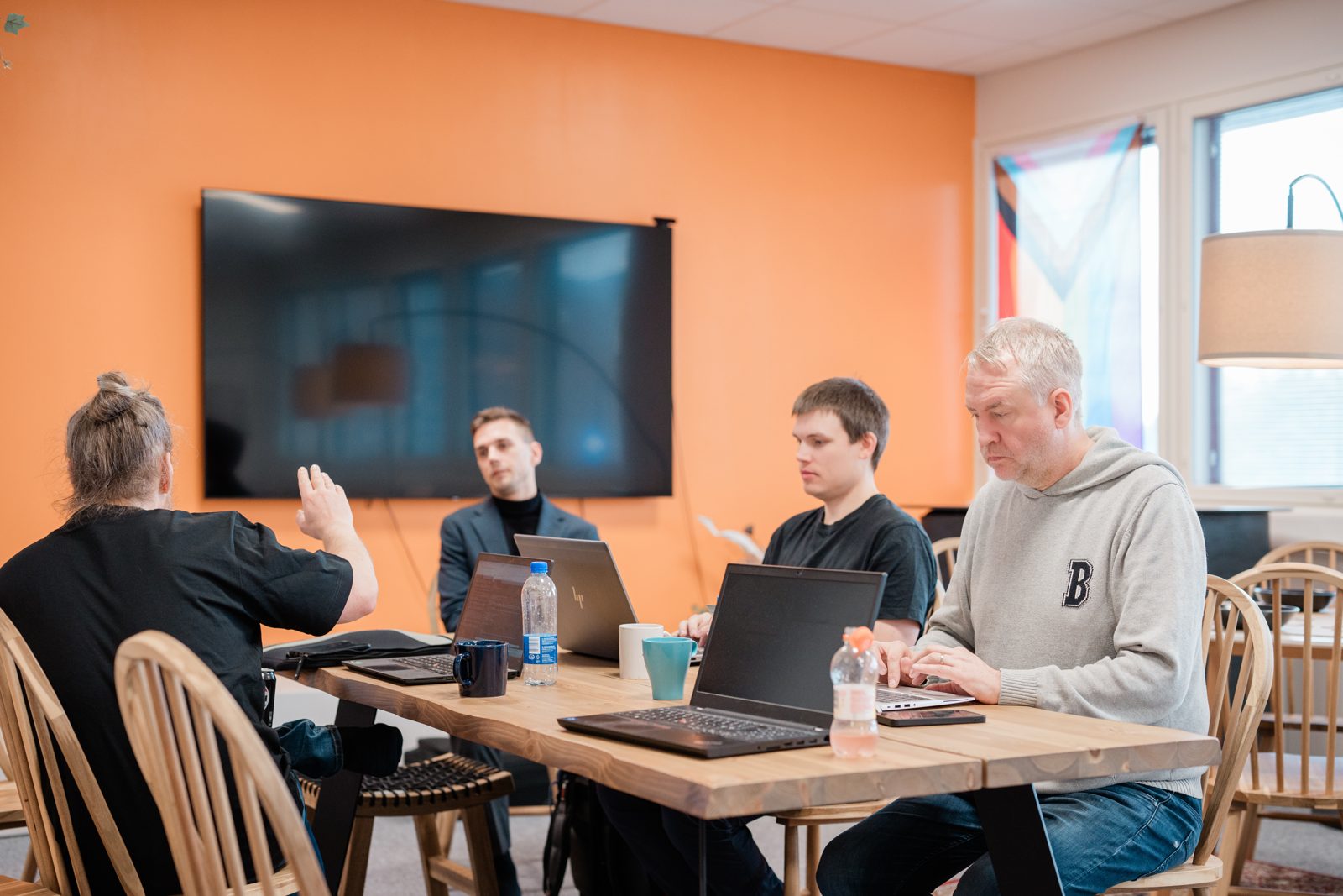 Viisi Sofokuksen asiantuntijaa palaverissa puupöydän ääressä kannettavien kanssa oranssin seinän edessä, yksi selittää / Five Sofokus experts in a meeting at a wooden table with laptops in front of an orange wall, one explaining with a hand gesture