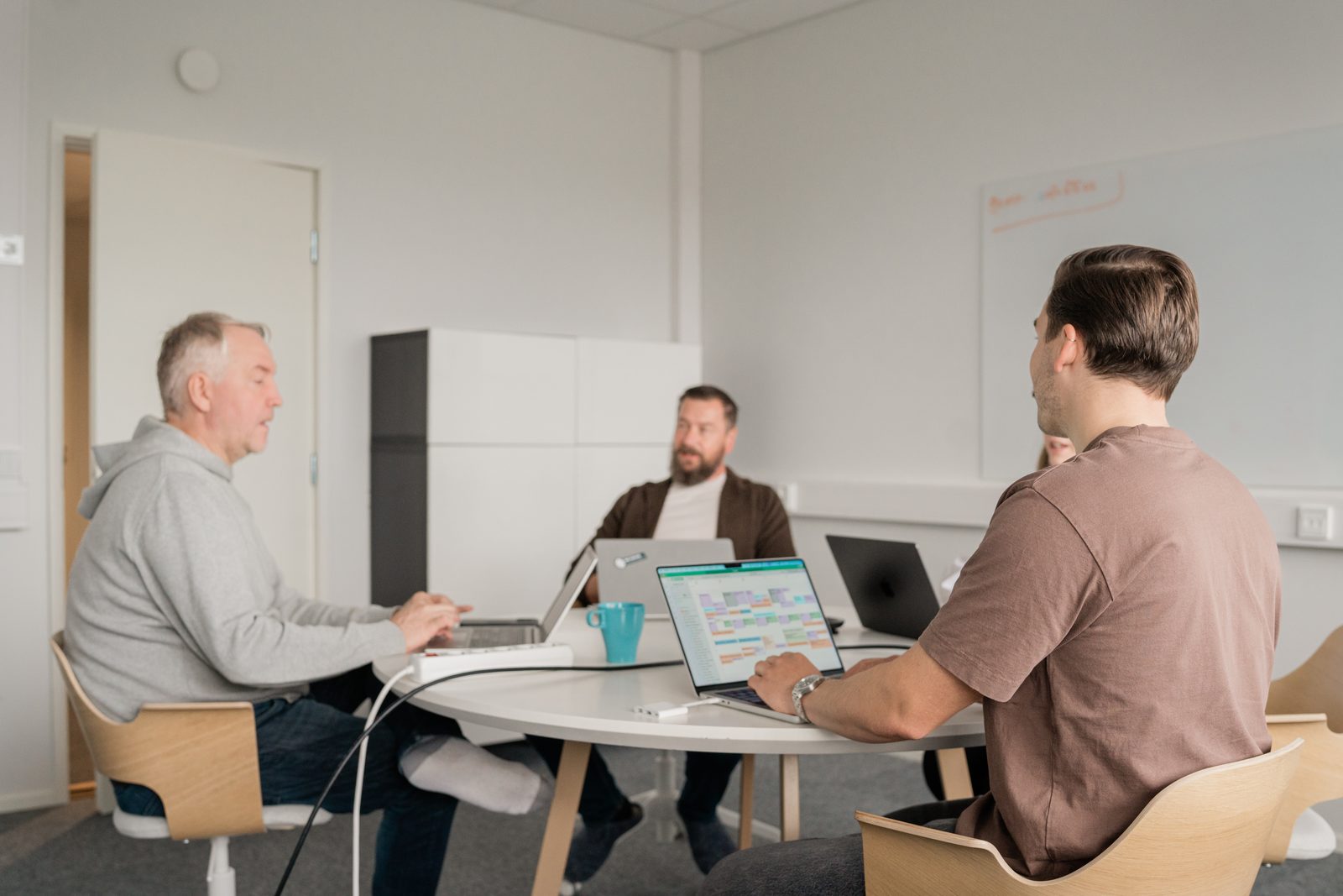 Kolme Sofokuksen asiantuntijaa palaverissa pyöreän pöydän ääressä kannettavat edessään valkoisessa neuvotteluhuoneessa / Three Sofokus experts in a meeting at a round table with laptops in front of them in a white meeting room