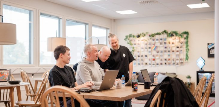 Istuva henkilö mustassa T-paidassa hymyilee, kun kollegat katsovat läppäriä – Digitaalisen liiketoiminnan sanasto