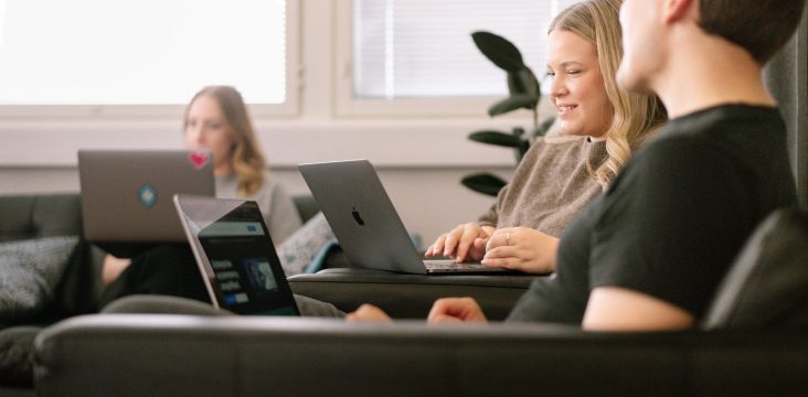 Hymyilevä henkilö kirjoittaa läppärillä, etualalla kollega mustassa T-paidassa – Siirtyminen Universal Analyticsista Google Analytics 4:ään: uudet ominaisuudet, tietosuojakeskeisyys ja hyödyt liiketoiminnalle