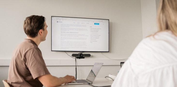 Henkilö läppärin ääressä katsoo seinänäyttöä neuvotteluhuoneessa – 10 syytä tehdä digiloikka tai aloittaa digiliuku