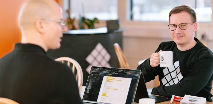 Hymyilevä henkilö Sofokus-collegepaidassa pitelee mukia pöydän ääressä – Missä sinun tietojasi säilytetään?
